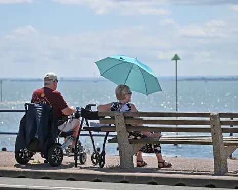 A man in a mobility scooter and a woman under a turquoise umbrella sit on a bench facing the sea