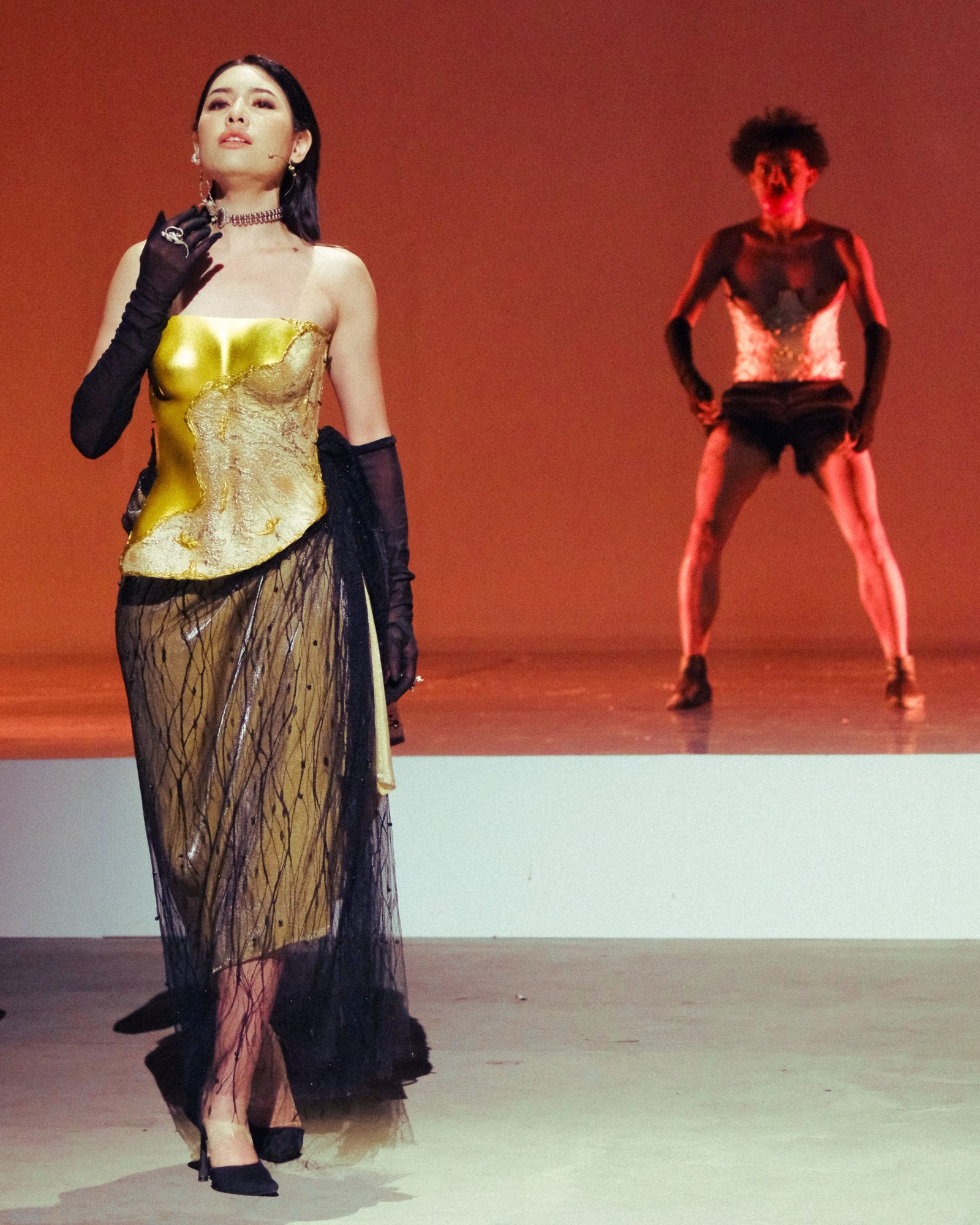 A performer in a gold and sheer black gown with long black gloves stands in the foreground on stage, while a shirtless dancer in a wide stance strikes a pose in dramatic red lighting in the background. Recipient of the Asian Cultural Council Grant cycle of 2026.