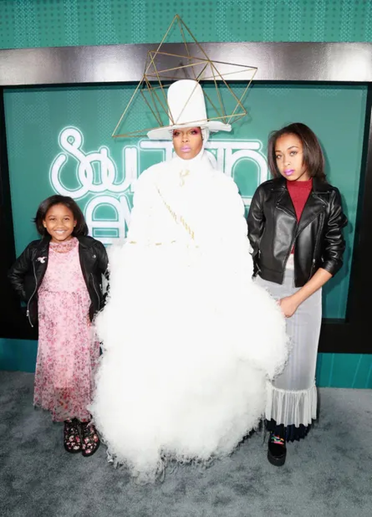 A woman in a white dress and hat stands with her daughters on either side of her at an awards show.