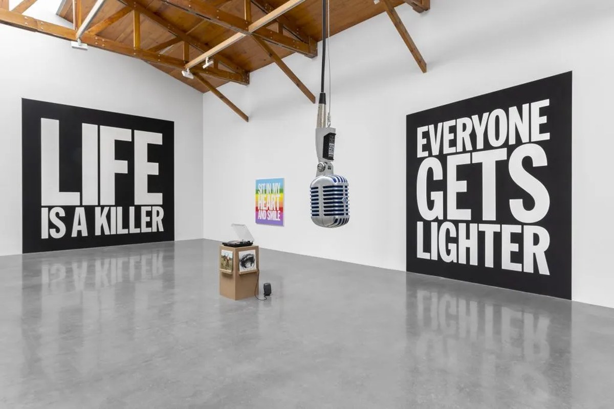 A photograph depicting a John Giorno exhibition staged in an airy, white-walled space with wooden ceilings featuring large black and white text-based works on the walls alongside smaller, more colorful ones, as well as a record player on a standalone plinth atop the concrete floor, and a metal microphone in the foreground hanging from the ceiling