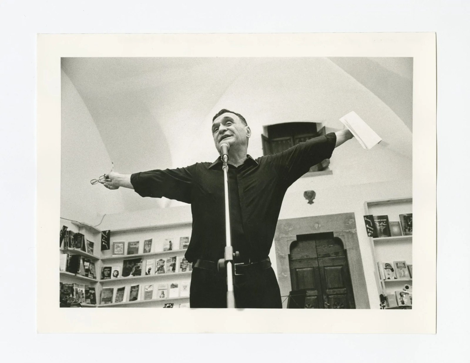 A black and white photograph looking up at John Giorno standing, arms-outstretched, at a microphone, wearing a black long sleeve shirt and black pants as he reads in a room lined with books