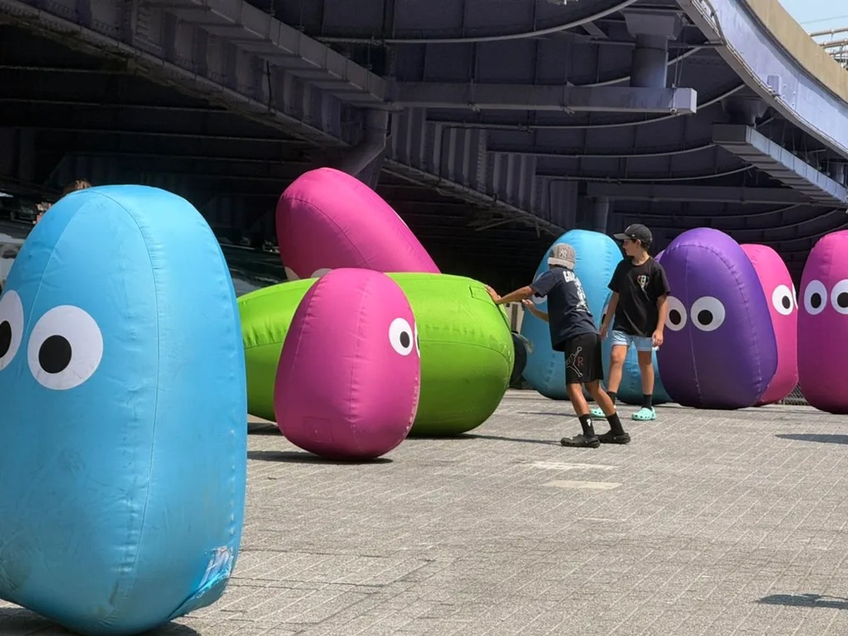 Rows of oversized inflatable blob-shaped creatures in blue, pink, green, and purple, each with large googly eyes, displayed outdoors on a paved plaza beneath a stadium or bridge structure. Two boys in caps interact with the inflatables.