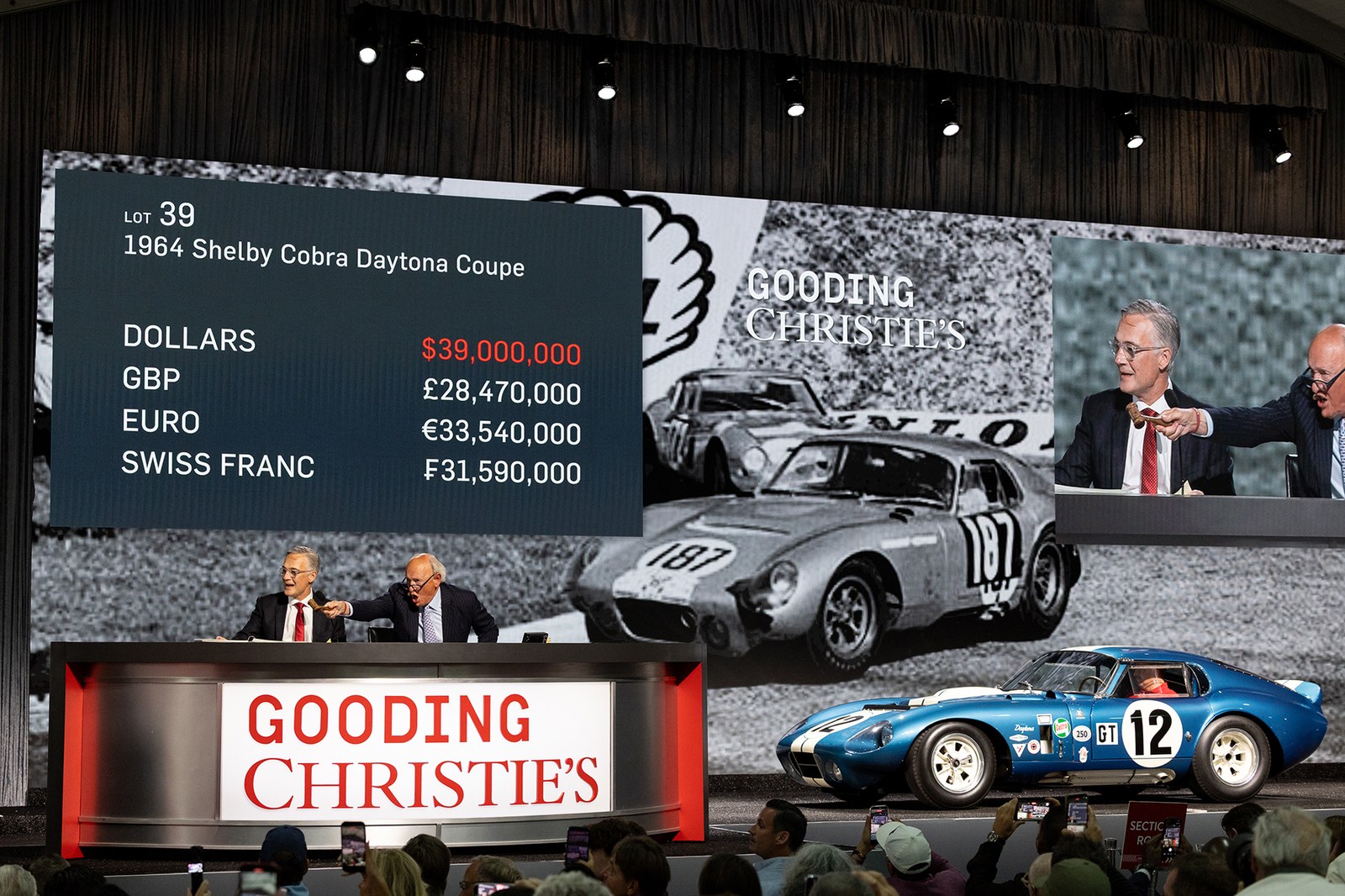 a photograph of an auction, we can see bids being taken, beside is a vintage blue racecar that is being sold