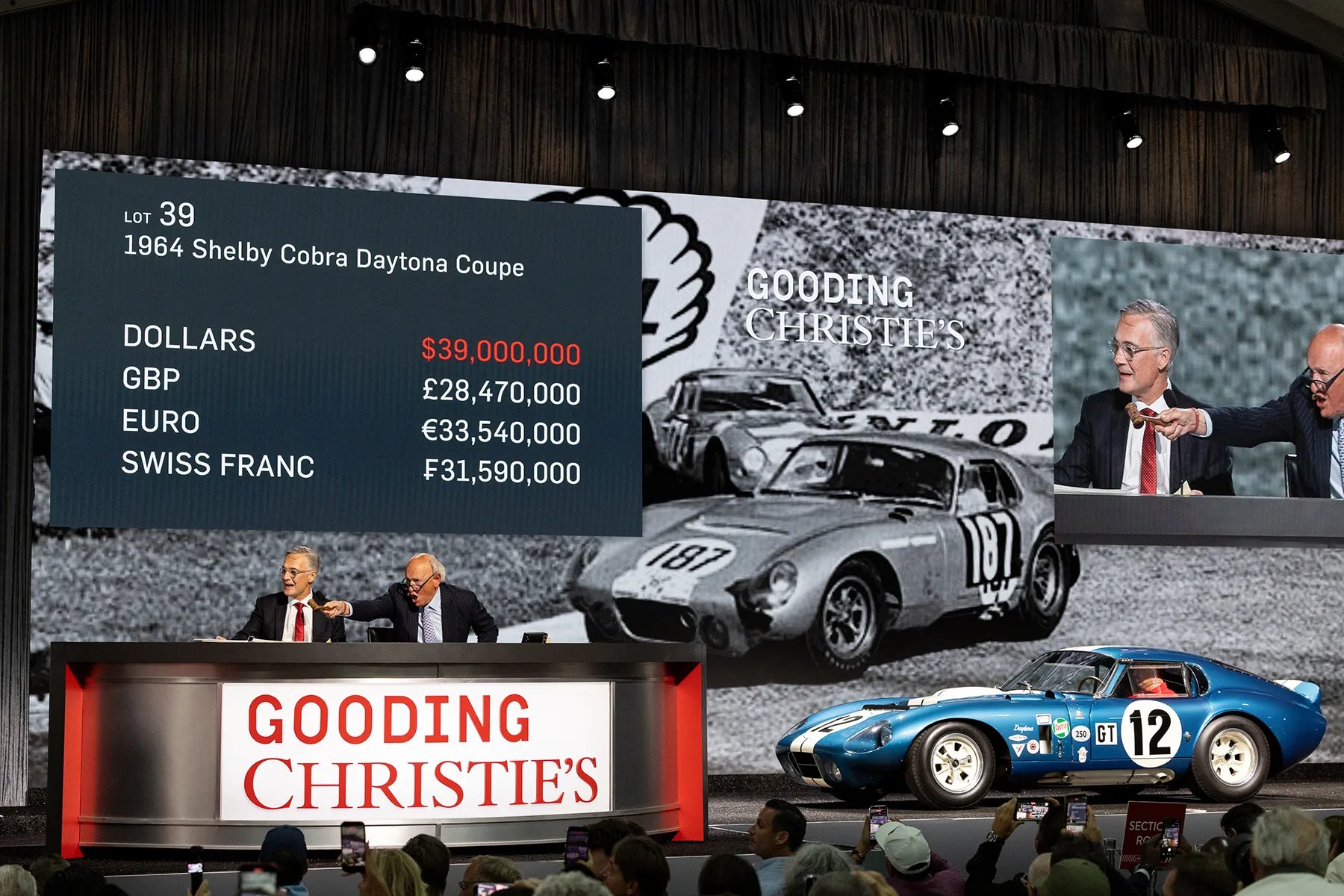 a photograph of an auction, we can see bids being taken, beside is a vintage blue racecar that is being sold