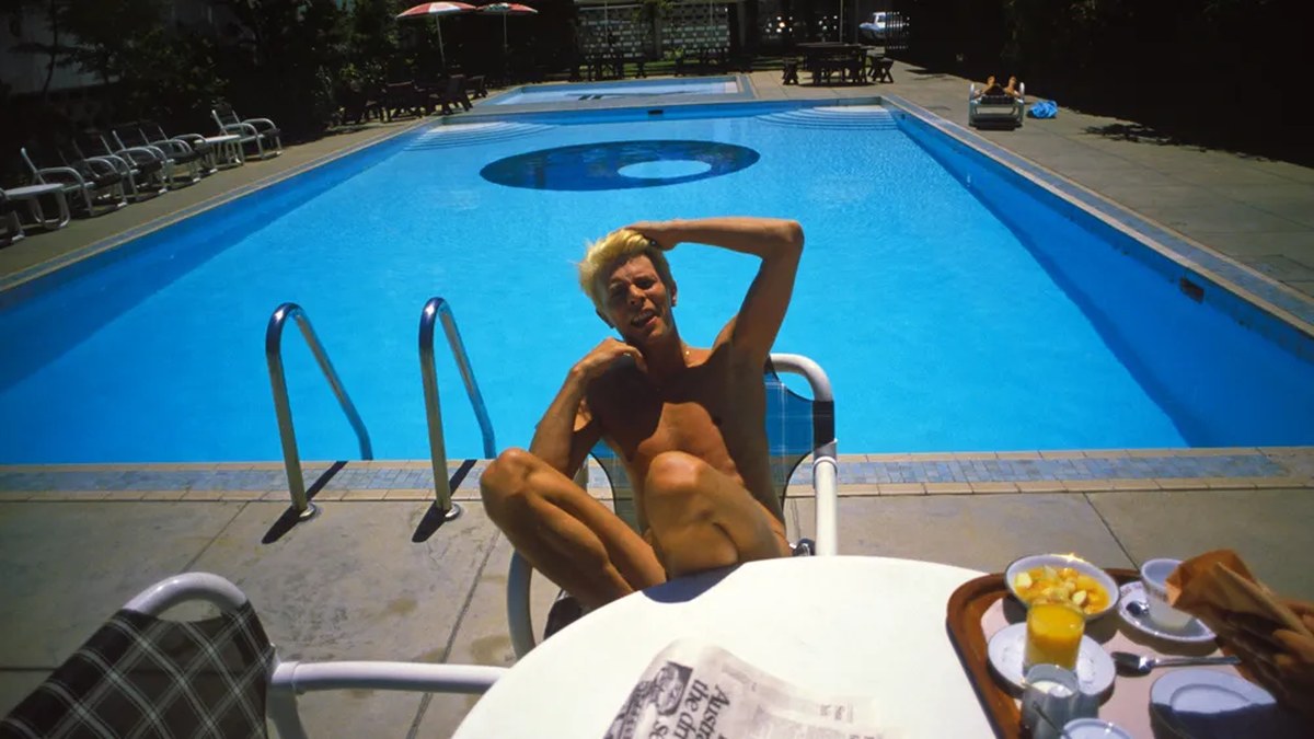 David Bowie relaxes poolside under bright Australian sun beside breakfast tray, shielding his eyes with one hand.
