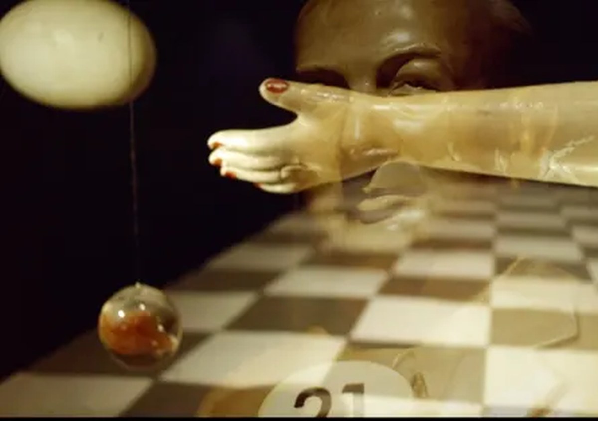 An artificial arm, with red-painted fingernails, throwing a glass apple over a chessboard.