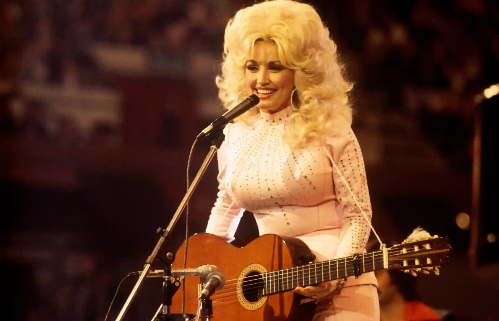A blond woman wearing a rhinestoned jumpsuit performs on stage with a guitar