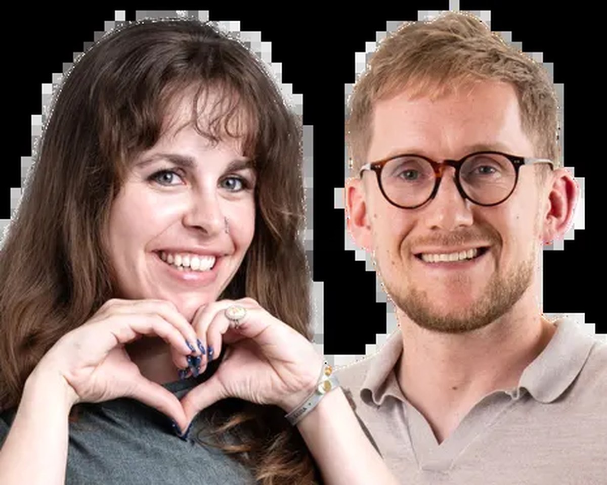 A woman making a heart shape with her hands stands beside a smiling man in glasses