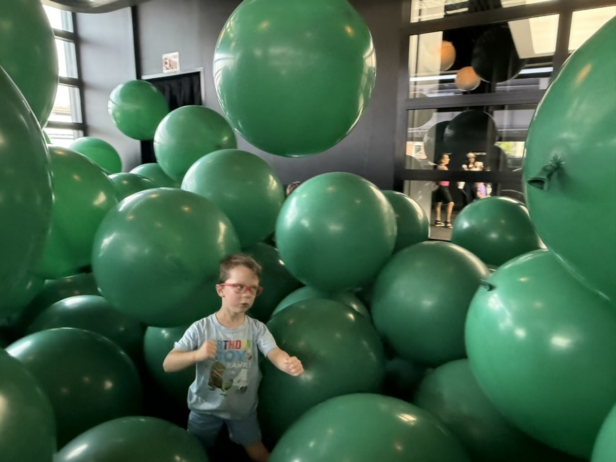 Installation view of Martin Creed's "Work No. 3883: Half the air in a given space" at the Balloon Museum, filling a room with green balloons; a young boy stands in the foreground among them, with onlookers visible through a window behind.