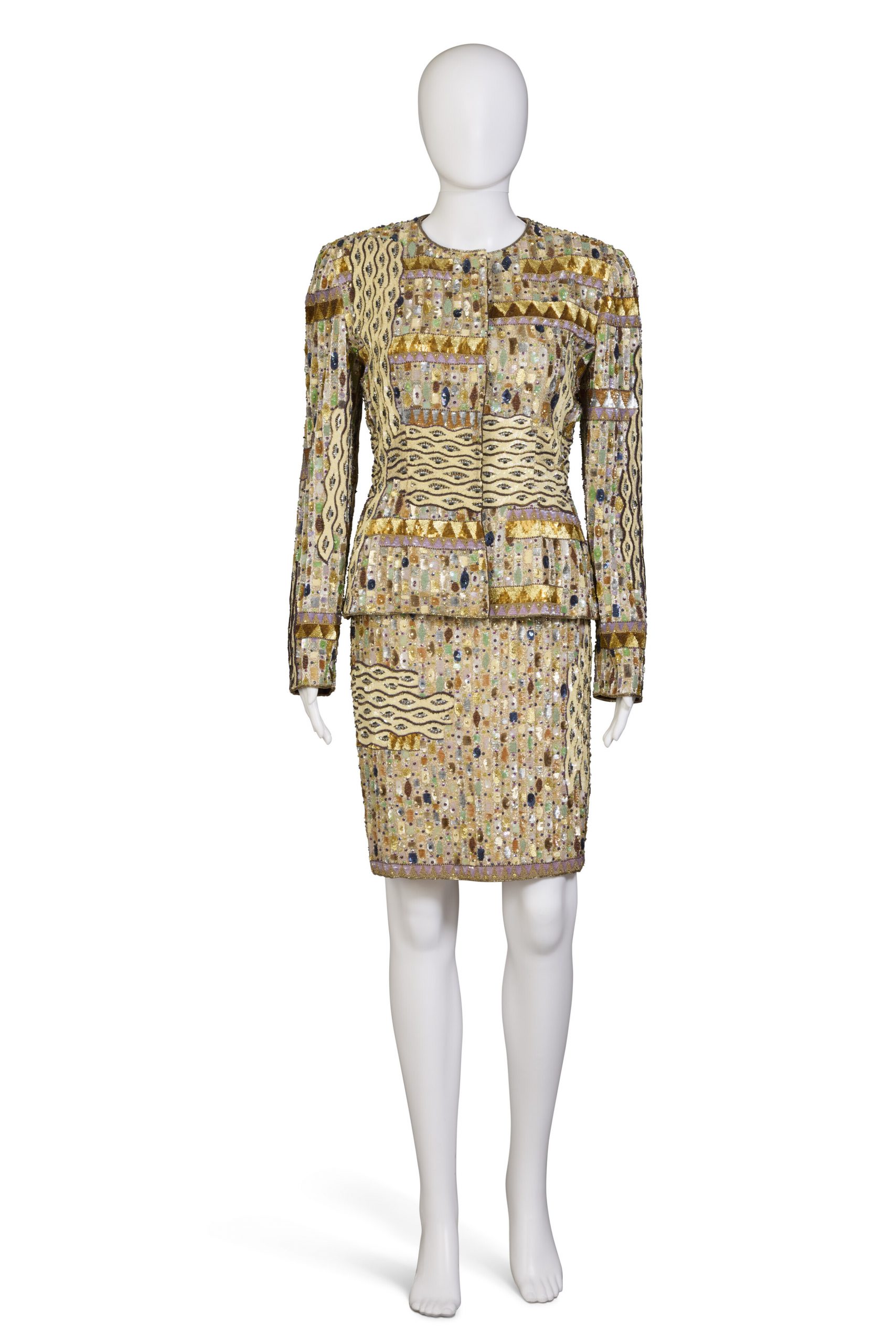 A photograph depicting a white mannequin wearing a gold knee-length skirt and jacket set by Bill Blass against an all-white background