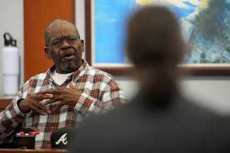 Man in flannel shirt sits in witness box, his hands over his chest and speaking, with blurry back of head in foreground.