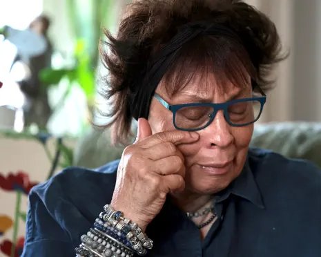 Joyce Thomas, 78, wipes her eye, wearing glasses, a headband and layered bracelets