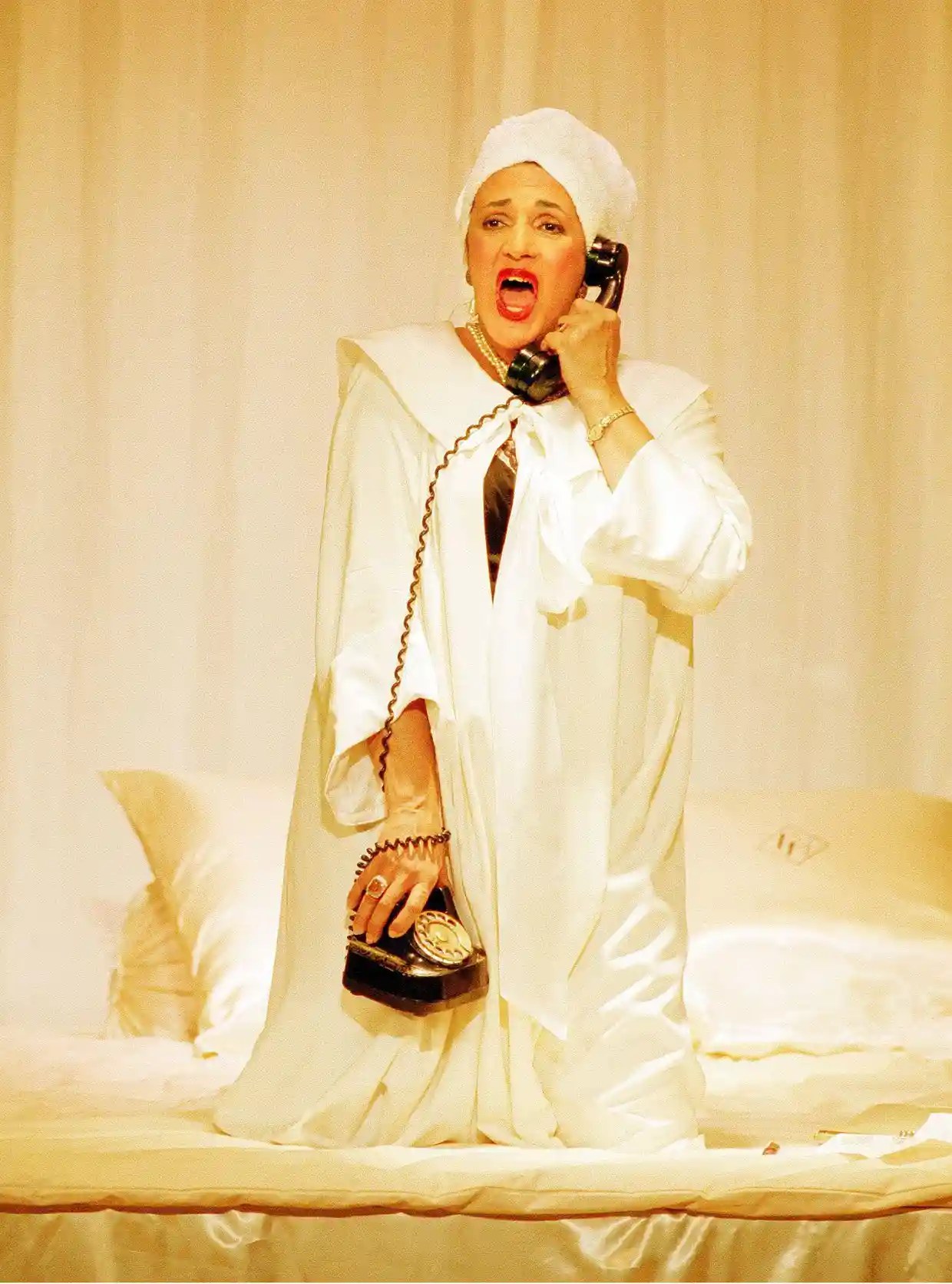 Jill Gomez on stage wearing a white dress and head wrap, holding a black telephone, while kneeling on a bed