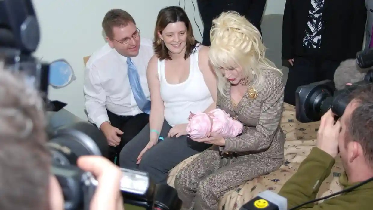 Dolly Parton holds newborn Emma wrapped in pink, beside her seated parents