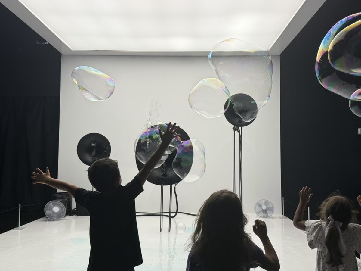 Children stand with arms raised in a dark, minimalist room, reaching toward large iridescent soap bubbles generated by fan-and-ring devices mounted on stands against a white wall.