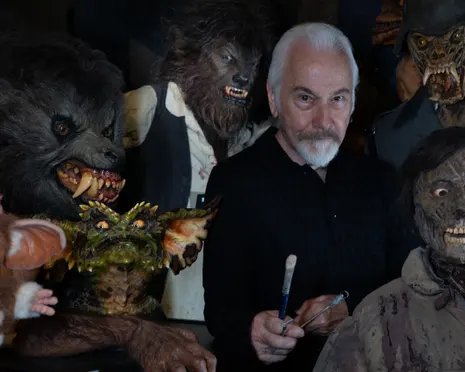 Rick Baker, holding paintbrushes, stands surrounded by his hairy monster and creature masks