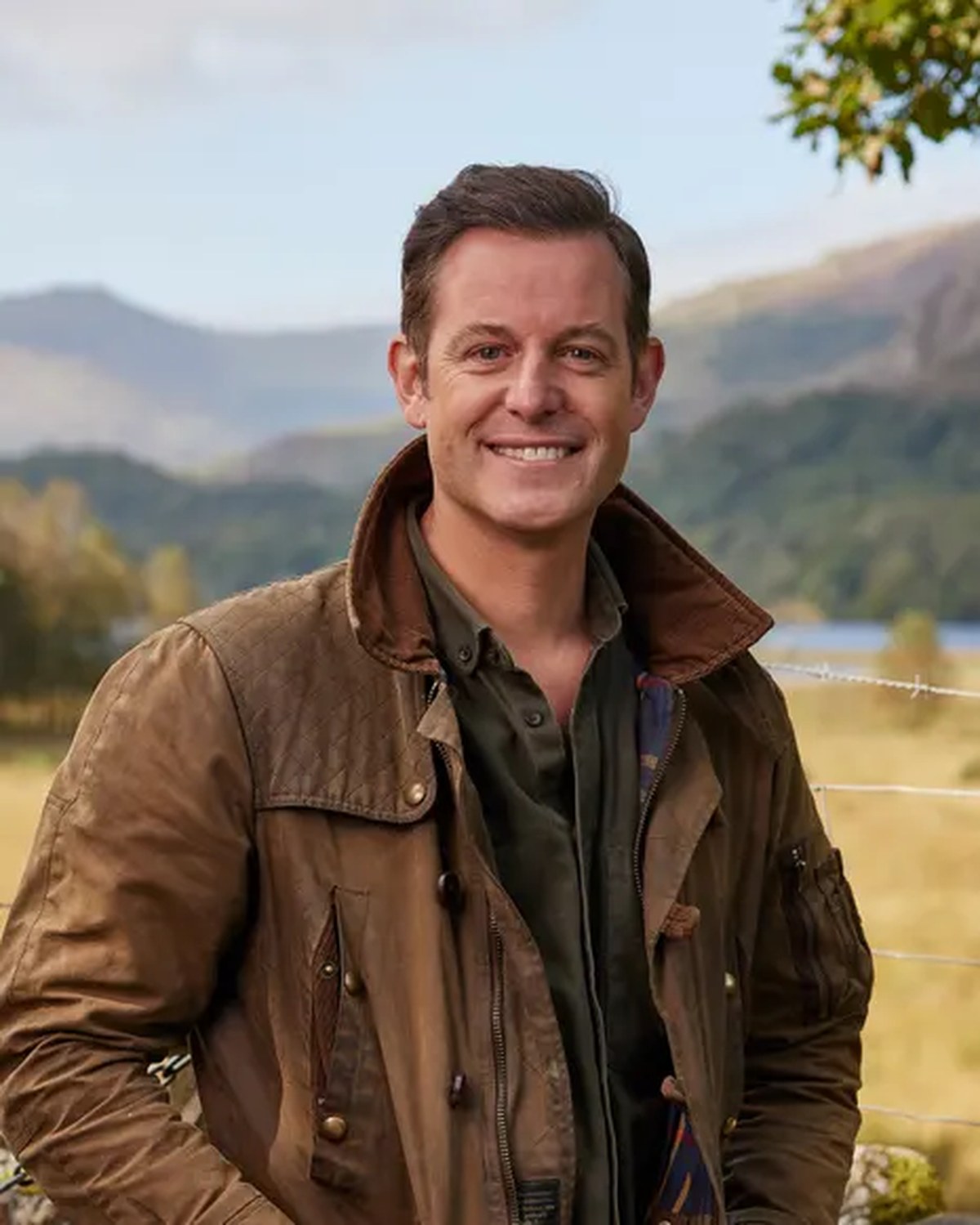 Matt Baker smiles in a brown jacket, standing outdoors with mountains and fields behind him
