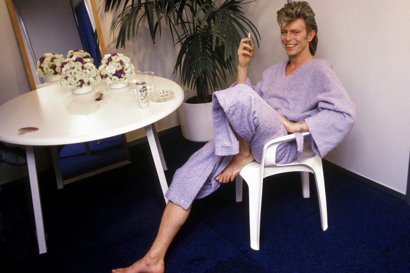 In a photo by Denis O'Regan, David Bowie relaxes backstage wearing a lavender robe, smiling while smoking beside flowers and mirrored dressing table.