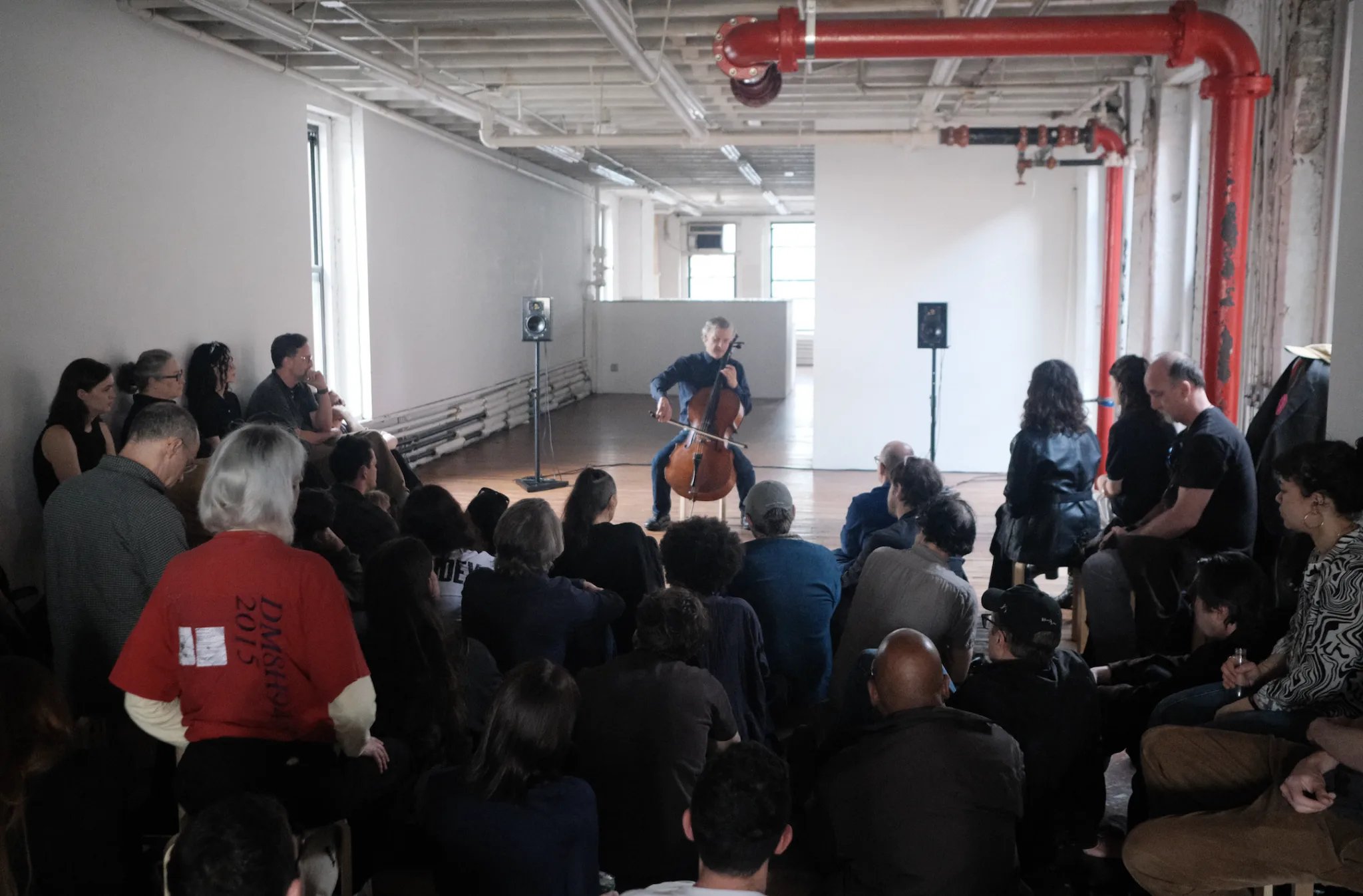 A photograph of a single cellist performing for a rapt crowd in a simple gray room with sunlight pouring in through the windows