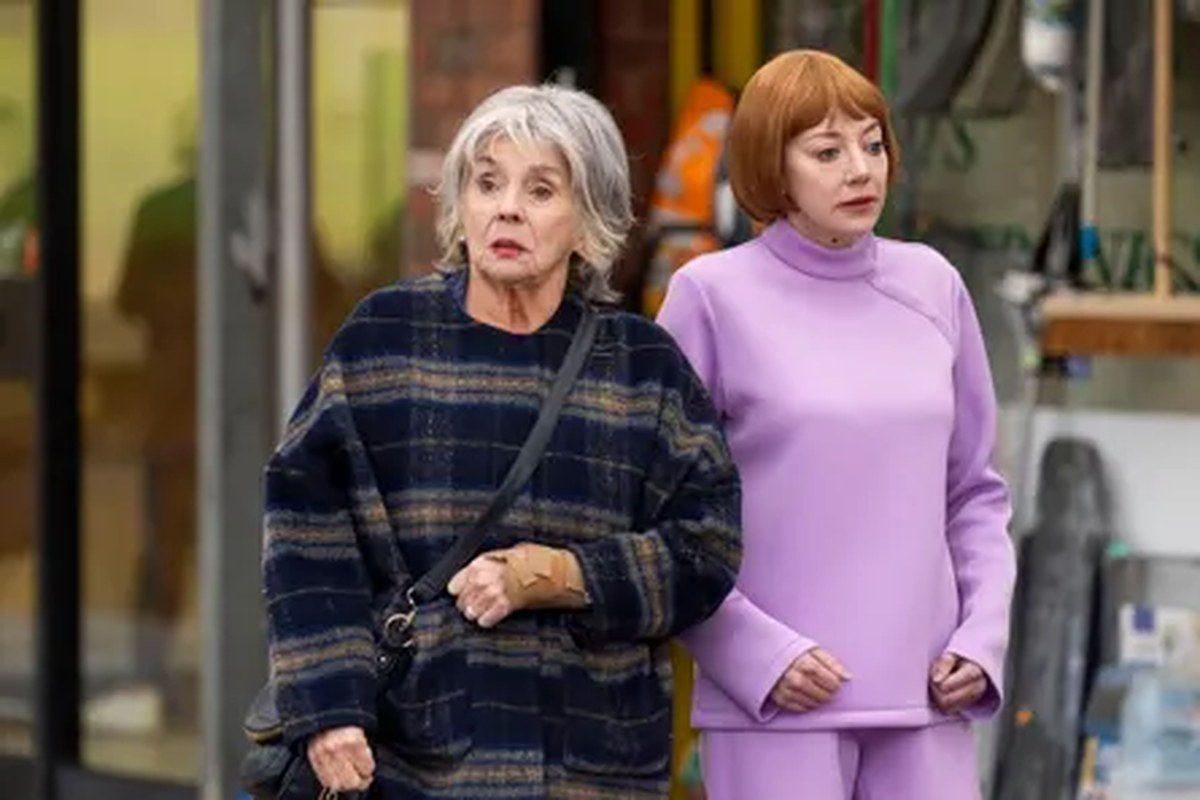 A woman with grey hair in a plaid coat standing next to a woman with red hair in a purple suit