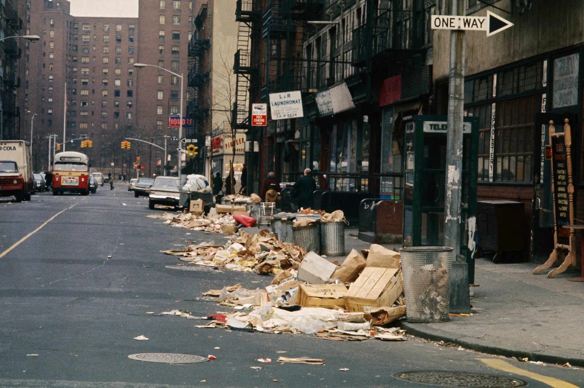 Archival color photograph of a New York City street lined with large trash piles along the curb. In the foreground, uncollected garbage — flattened cardboard boxes, torn paper bags, wooden crates, and loose debris — spills from overflowing metal trash cans onto the sidewalk and into the gutter for most of a block. A green telephone booth marked "TELEPHONE" and a "ONE WAY" street sign stand near the curb. Behind the trash, a row of storefronts includes "L & R LAUNDROMAT" and a furniture/upholstery shop, with tenement buildings and fire escapes rising above. A "BUS STOP NO STANDING" sign and a "LIQUORS" sign are visible further down the block. A vintage red-and-cream city bus, a boxy delivery truck, and several parked cars line the street, with a few pedestrians walking near the shops. In the far background, a tall brick high-rise apartment building overlooks the scene under an overcast sky.