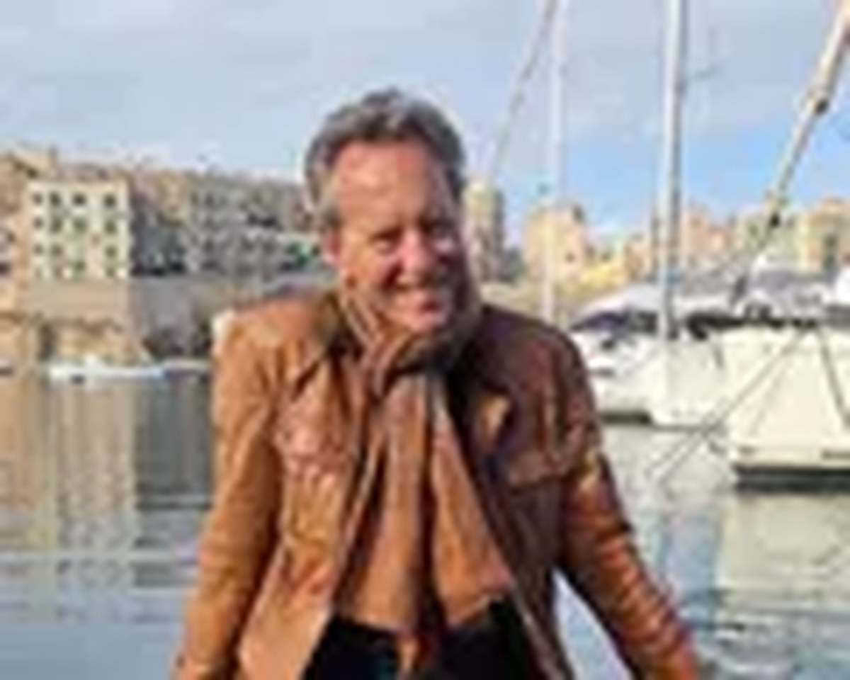 A man with a big grin on his face wearing a tan leather jacket and scarf sitting in front Malta’s Grand Harbour