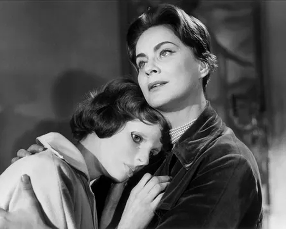 A black and white image of a girl with a mask on, clasping on to a woman in a black jacket