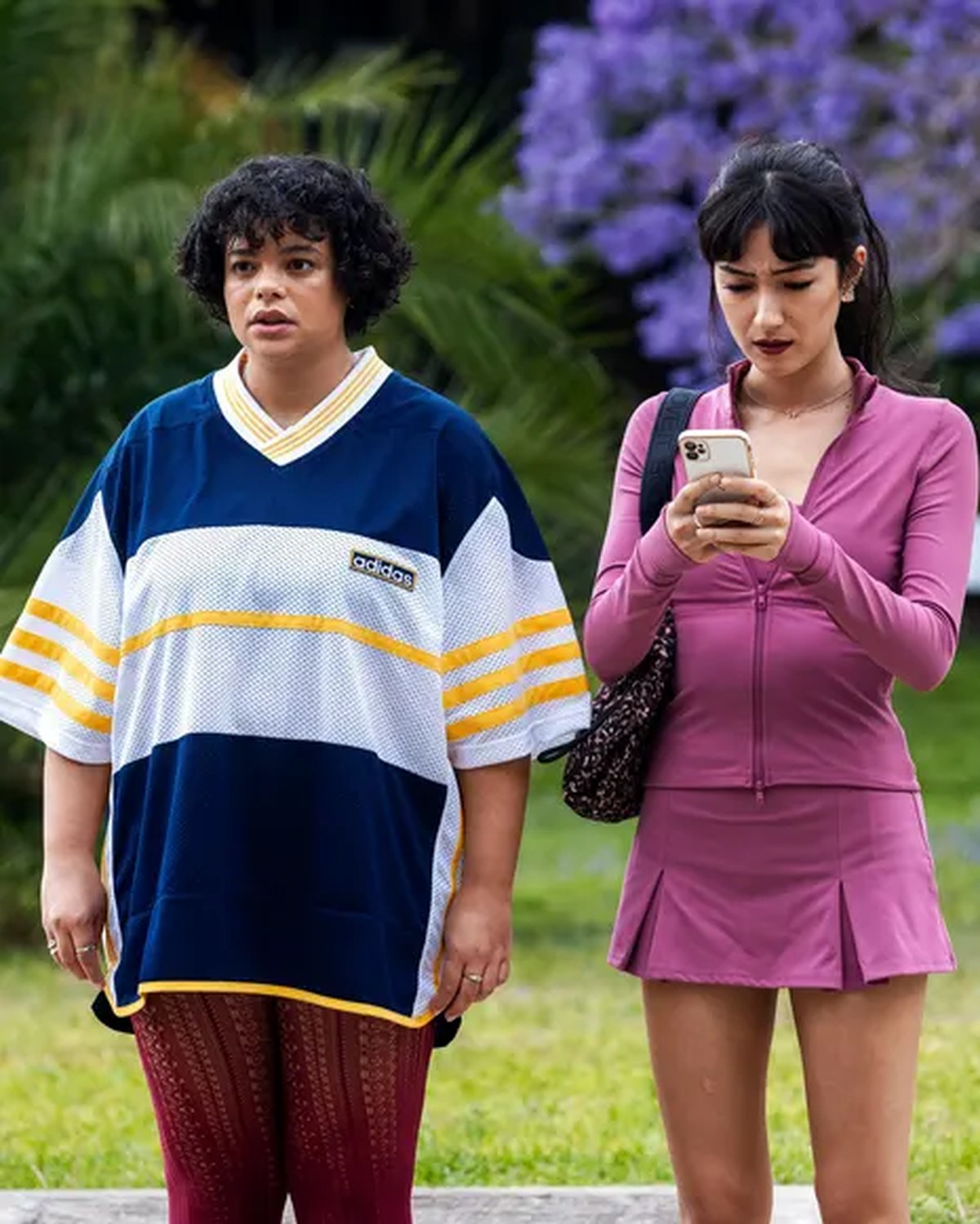 Two women - one in a chic pink suit and another in an oversized blue and white top and brown trousers standing in green area with trees and flowers behind them
