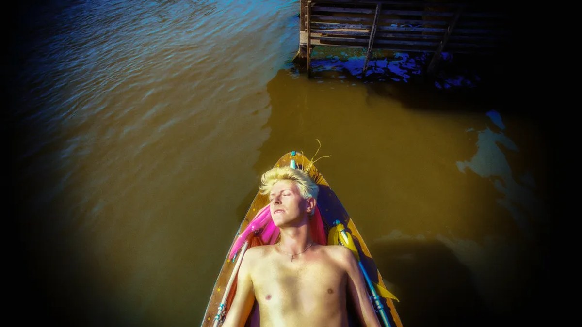 In a Denis O'Regan photo, David Bowie sunbathes shirtless inside a narrow canoe floating beside a dock on calm brown river water peacefully.