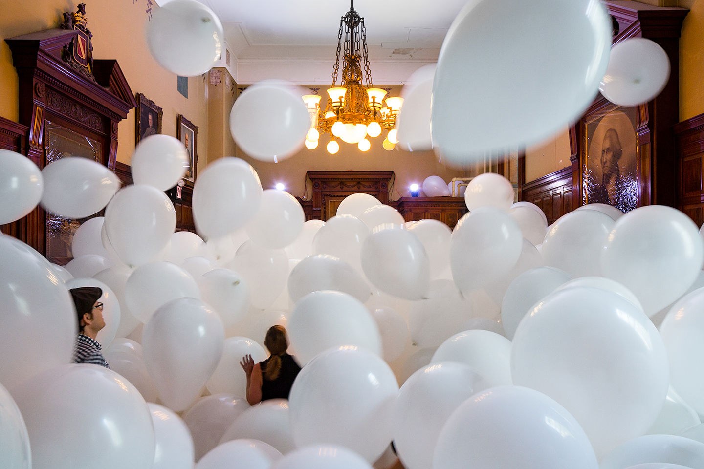 Installation view of Martin Creed's "Work No. 3883: Half the air in a given space," filling an ornate, wood-paneled room with white balloons floor to ceiling beneath a gilded chandelier and framed portraits; two visitors are visible amid the balloons.