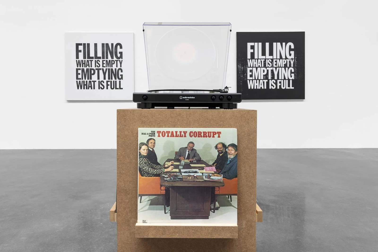 A photograph depicting a record table atop a wooden plinth displaying a record on a ledge on its front face, with two black and white text-based artworks hanging on the white wall behind it