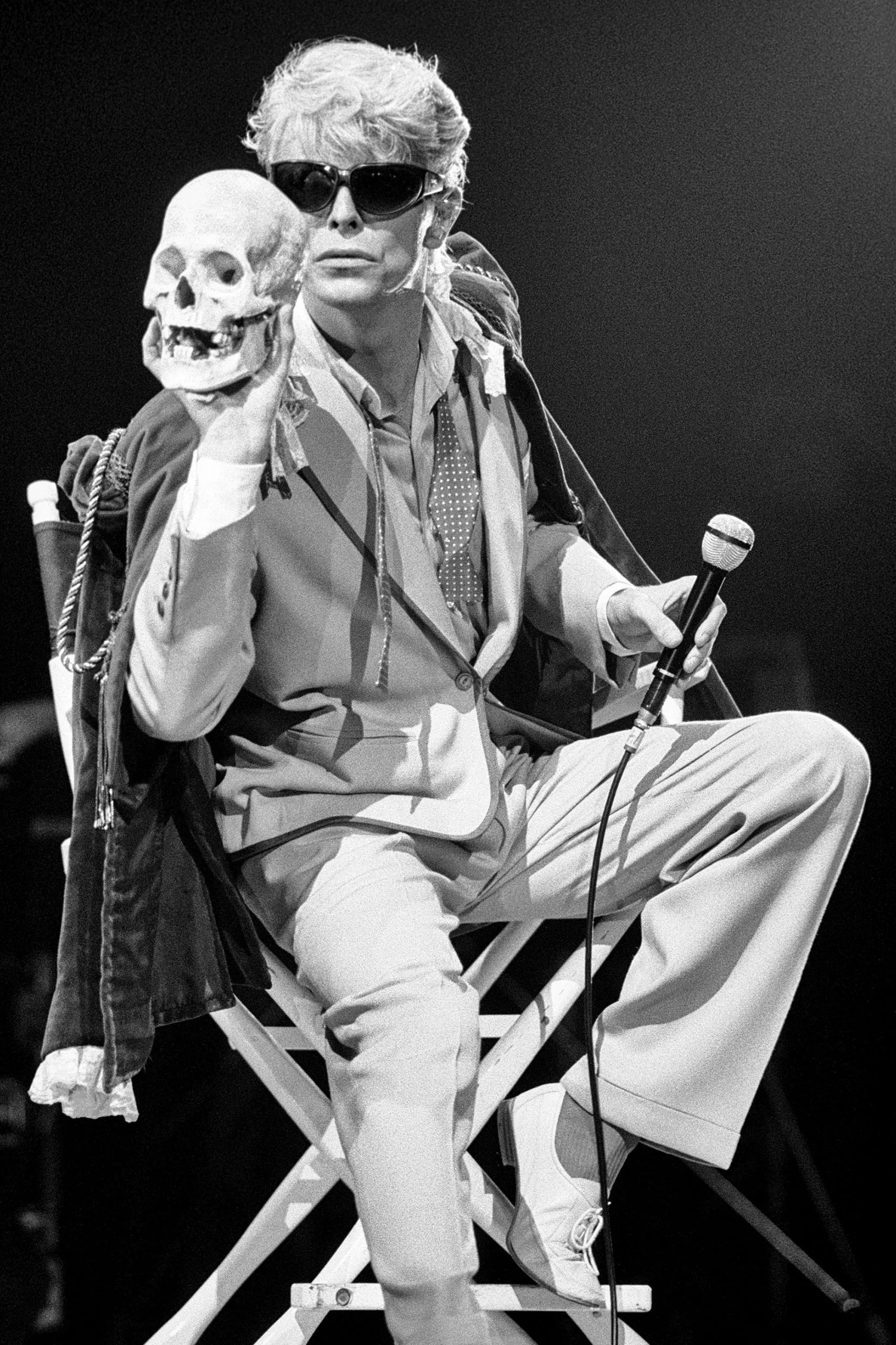 David Bowie, wearing sunglasses, holds a human skull and microphone while seated during theatrical stage performance.