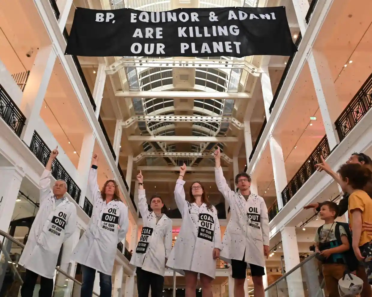Five activists in white lab coats with ‘Coal out of our museums’ badges line up in the Science Museum’s energy hall under a banner that reads ‘BP, Equinor and Adani are killing our planet’