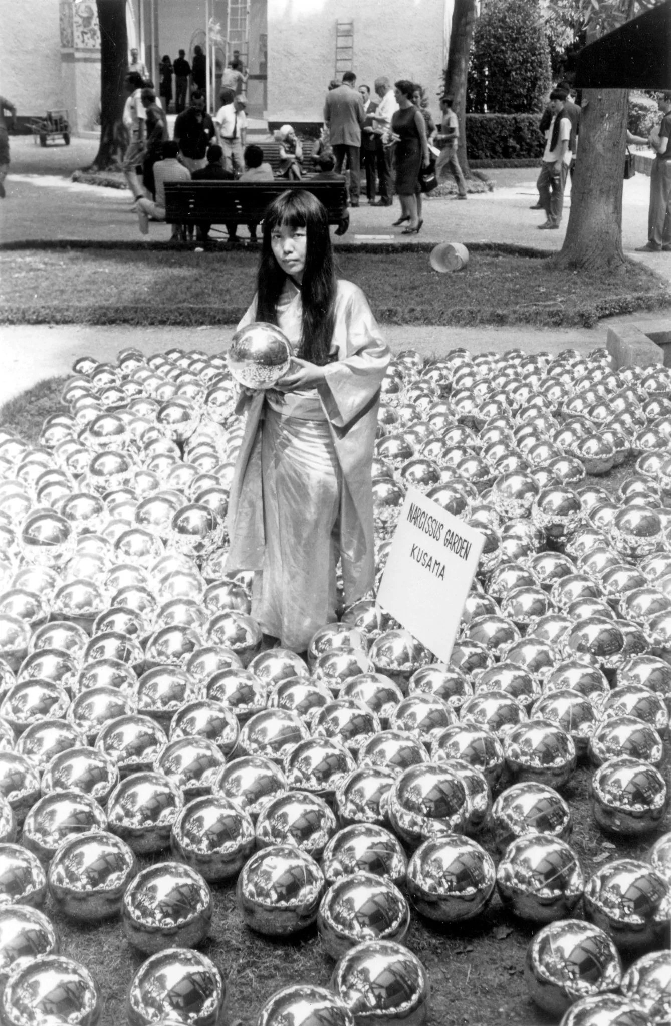 a black and white photograph in which a woman holds a shiny looking ball while standing in a field of similar balls
