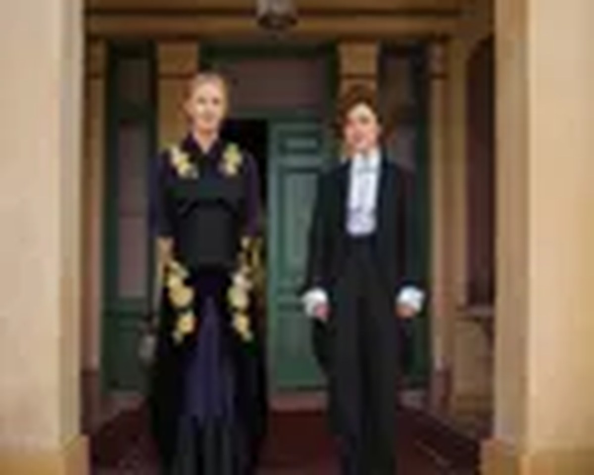 In the doorway of an elegant house a woman in a long embroidered dress stands with her hands behind her arms next to a young woman with her hair piled on her head wearing a tailored suit, trousers and bowtie