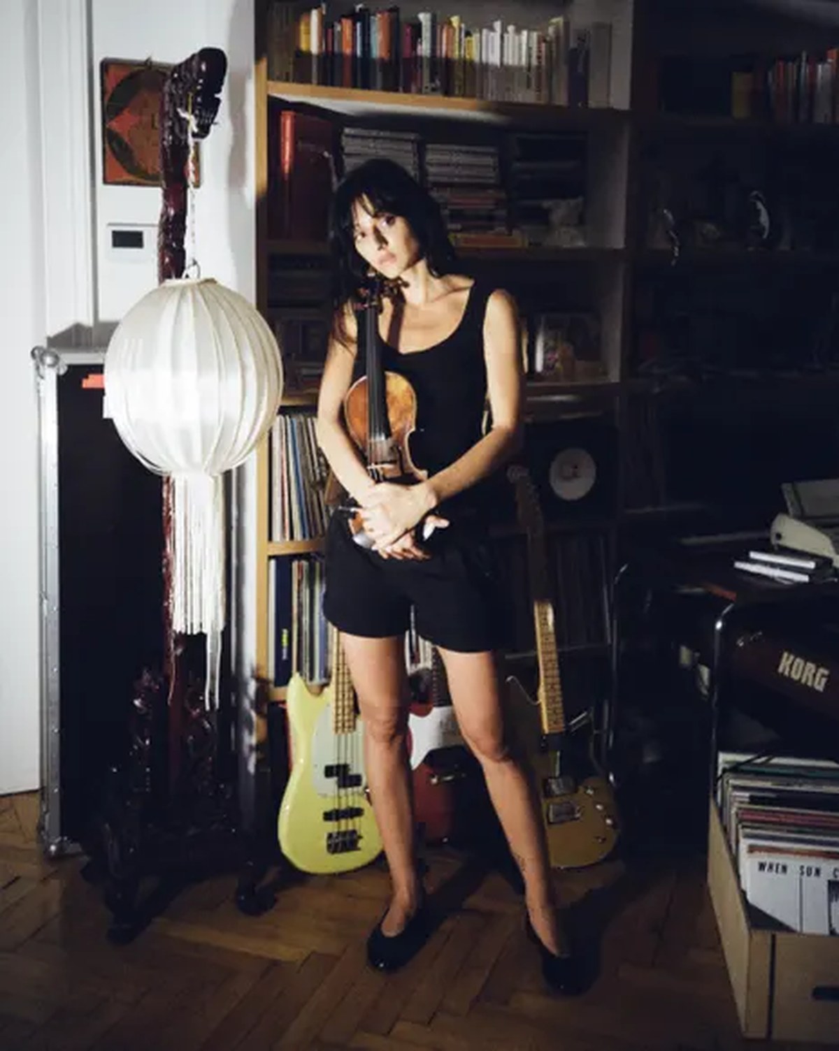 Singer Sofie Royer holds a violin, standing before a bookshelf and paper lantern lamp