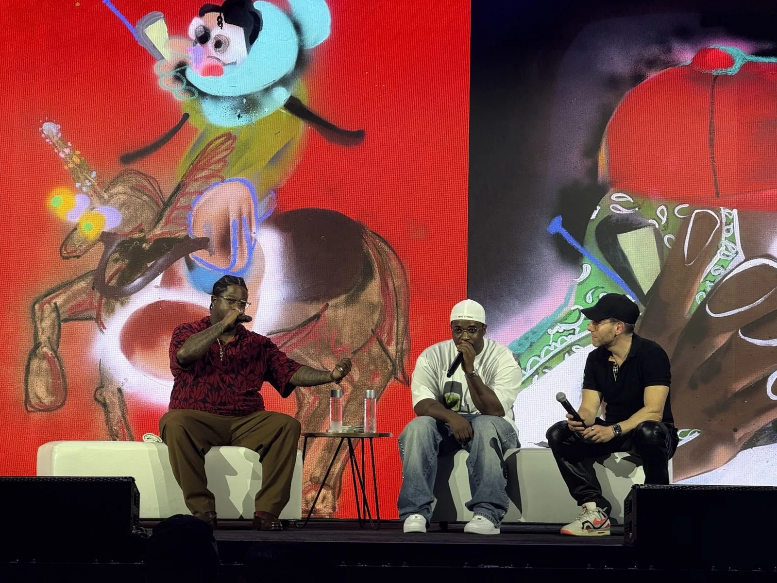 Three panelists seated on a stage holding microphones in front of a large screen displaying colorful, expressive cartoon-style artwork on a red background. The man on the left wears a red patterned shirt and brown pants, the man in the center wears a white t-shirt, white cap, and jeans, and the man on the right wears a black polo, black pants, and a black cap. Example of type of programming to be featured at InScope 2026.