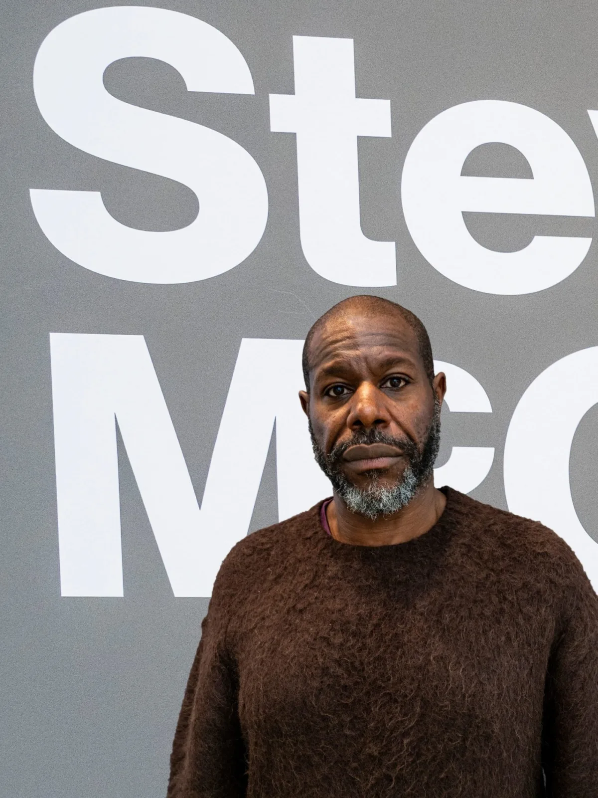 a man in a brown jumper poses in front of some white writing printed onto a grey ground, we only see him from the chest up