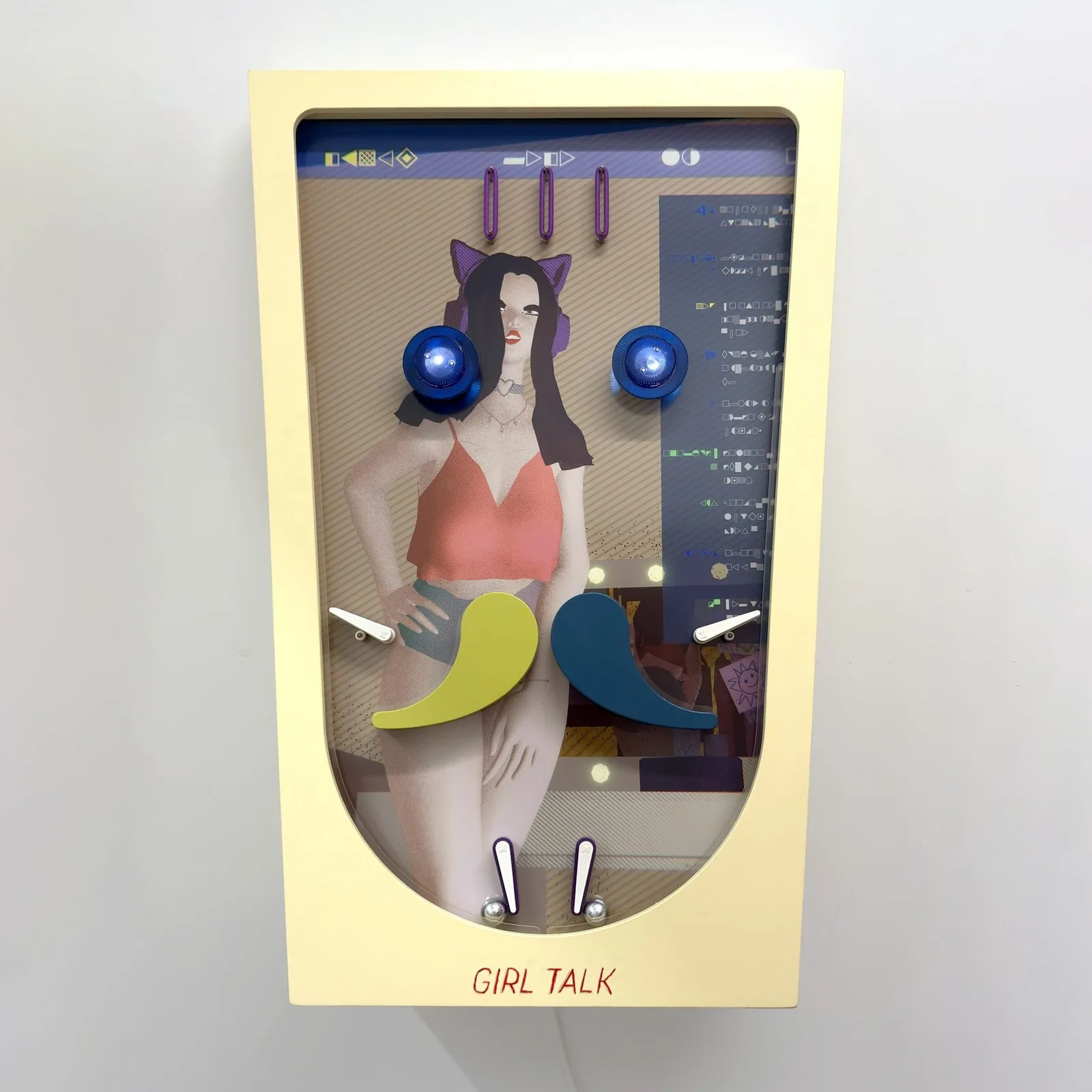 Tall, pale yellow wall-mounted shadowbox with a rounded bottom and the title “Girl Talk” at the base. Inside, an illustration of a young woman with long dark hair, purple cat-ear headphones, a choker, a coral crop top and shorts, standing with one hand on her hip. Behind her is a computer-screen interface with rows of abstract symbols. Pinball parts sit over the image: two glowing blue bumpers by her face, two large curved yellow-green and teal paddles at hip level, small white flippers at the sides, and two more flippers with silver balls at the bottom.