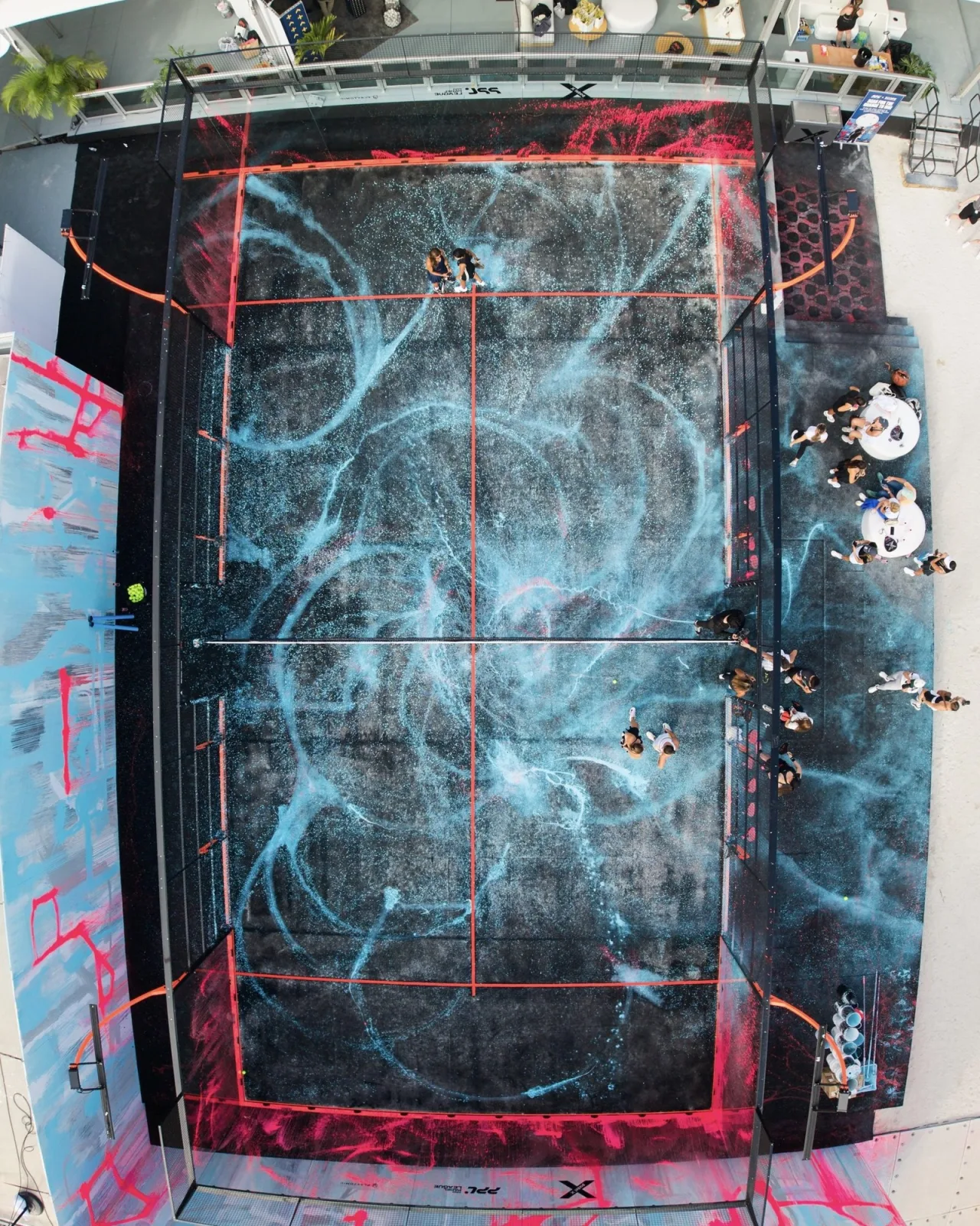 Aerial view of an artistically painted pickleball court in Miami, its black surface covered in sweeping, splattered blue and pink paint strokes that evoke a cosmic or wave-like pattern, bordered by orange lines marking the playing area. Several players are visible mid-game on the court, with spectators and tables gathered along the right side. Example of type of project to be featured at InScope 2026.