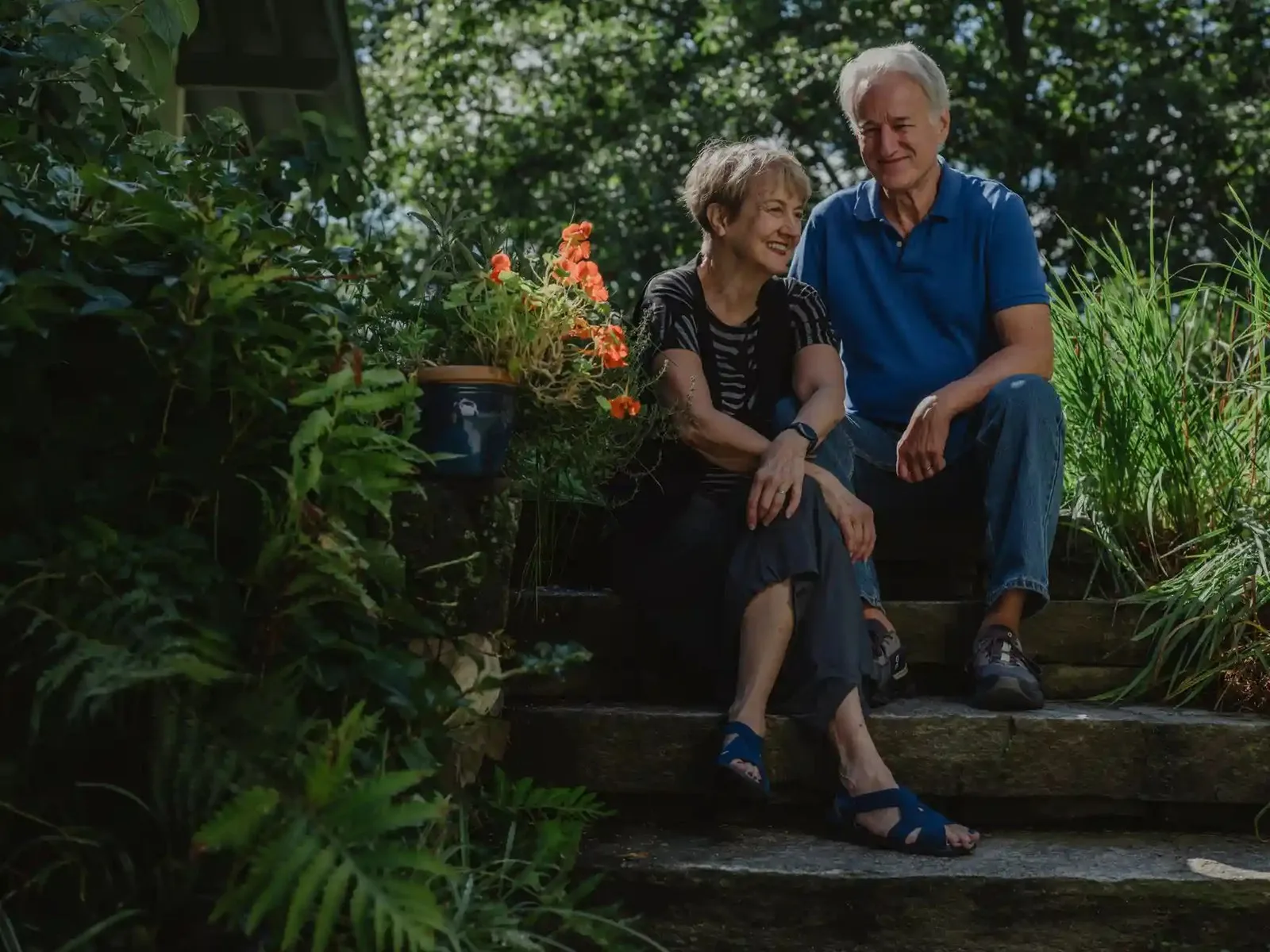 A man in a blue top and a woman in a stripy top and a black vest, sitting on steps in a garden