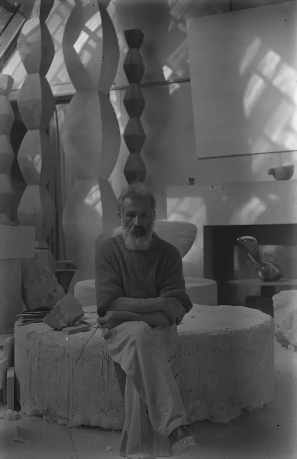 A bearded man sits in an artist’s studio surrounded by tall cylindrical sculptures and a sculpted cup on the mantel of a fireplace.