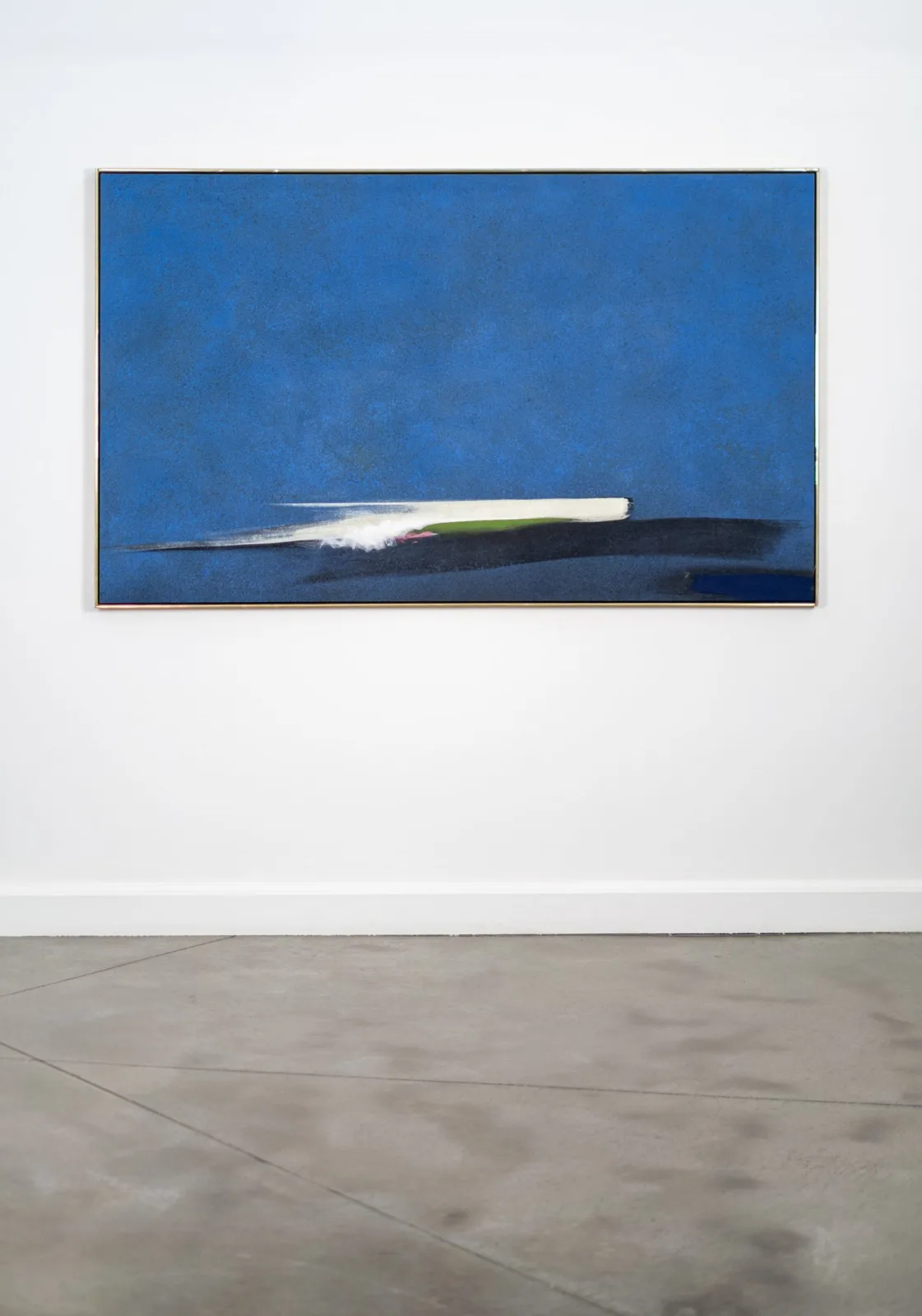 Large horizontal abstract painting in a thin brass frame, hanging on a white gallery wall above a concrete floor. A deep, speckled blue field fills most of the canvas. Low across the bottom, a streak of cream-white paint with a small patch of green and a touch of pink sweeps from left to right over a dark navy-black band, like a boat or wave skimming the horizon. Included in the Oeno Gallery exhibition Constructing Space