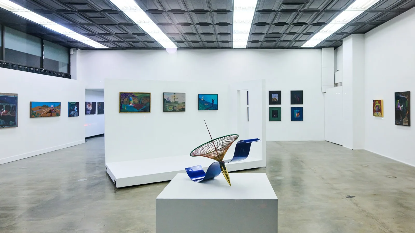 An art gallery interior in a Veronica Mishaan Bogotá penthouse, displaying several framed paintings on white walls and a large, abstract metal sculpture on a pedestal in the foreground.