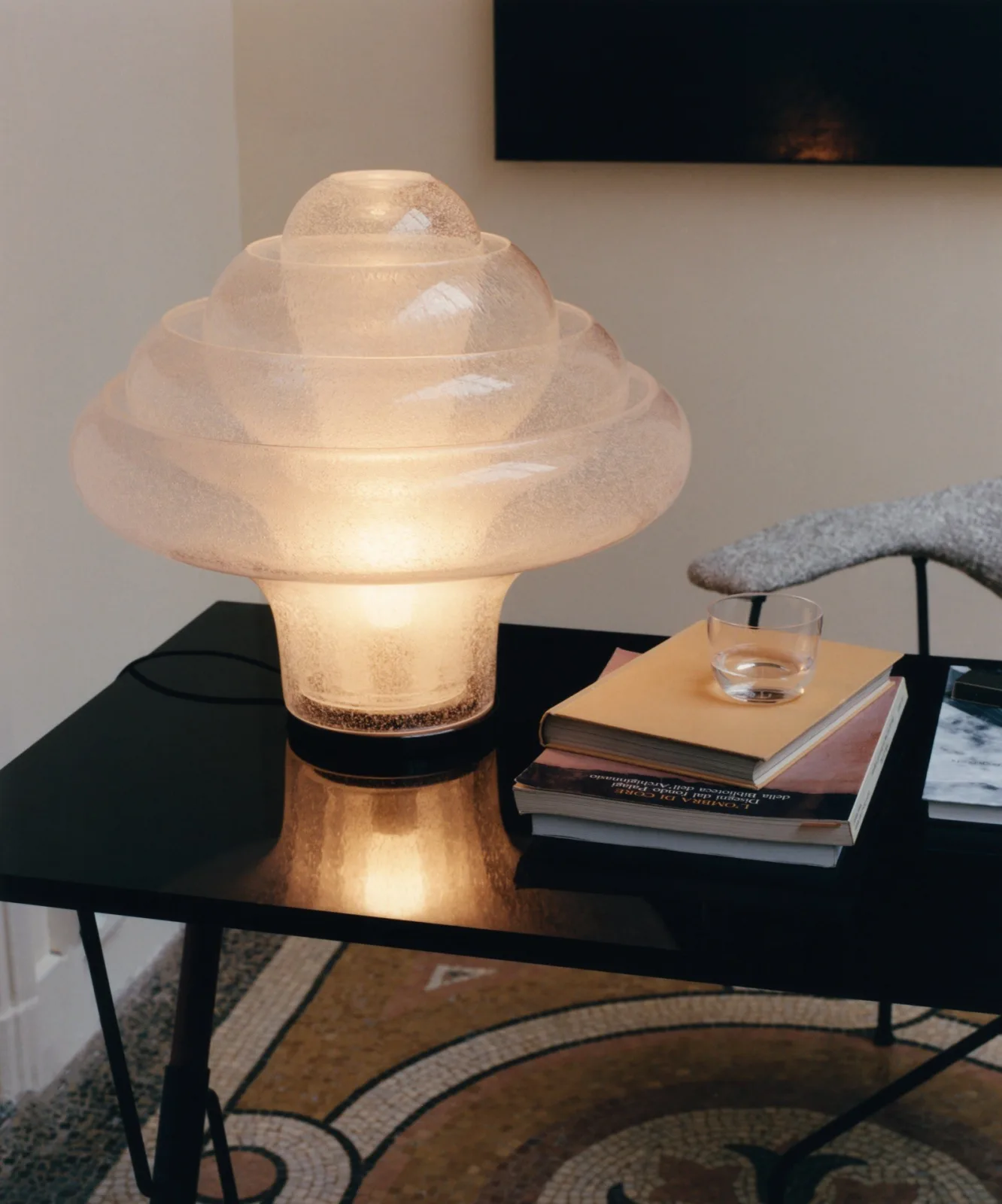 A tiered, textured glass table lamp glowing on a black side table next to a stack of books and a small glass of water.