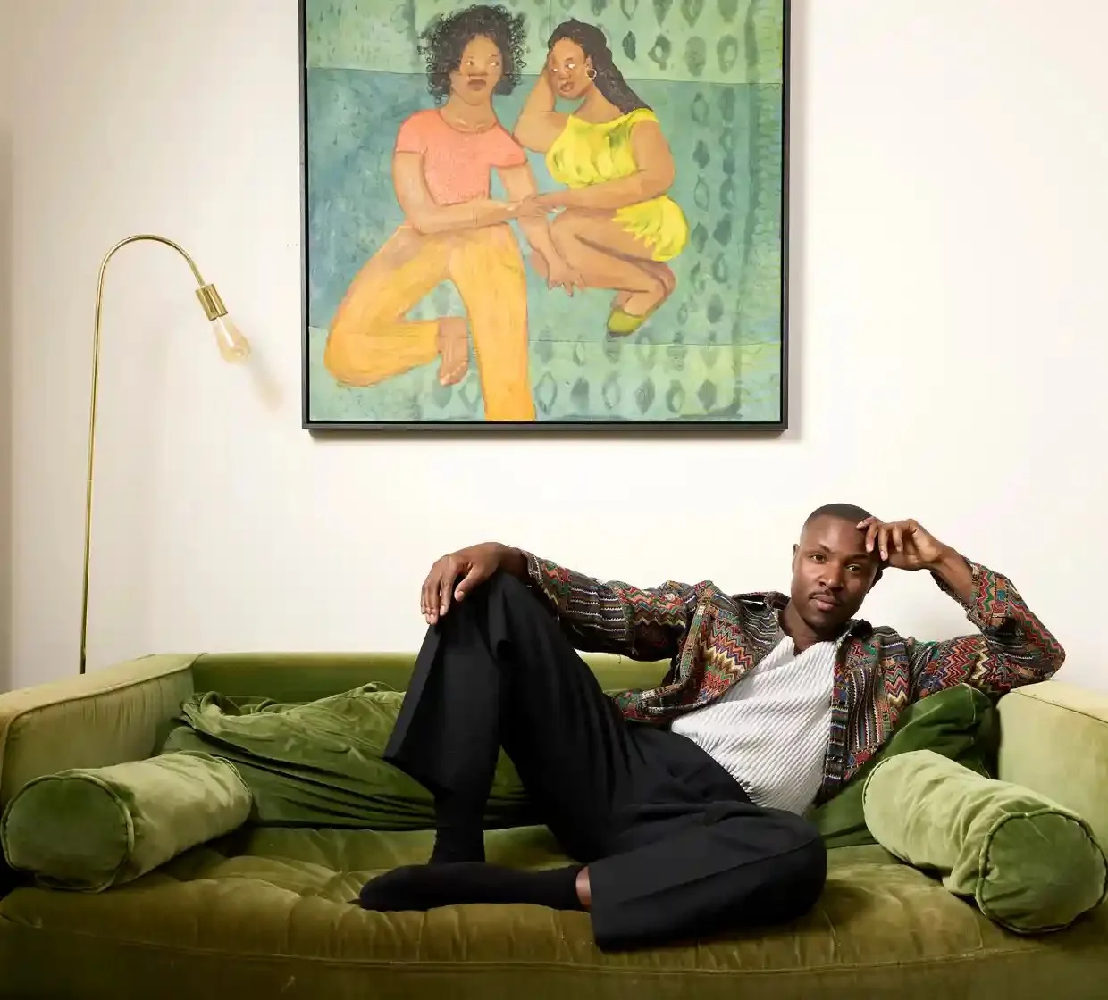 He sits on a green velvet sofa beneath an artwork showing two black women holding hands