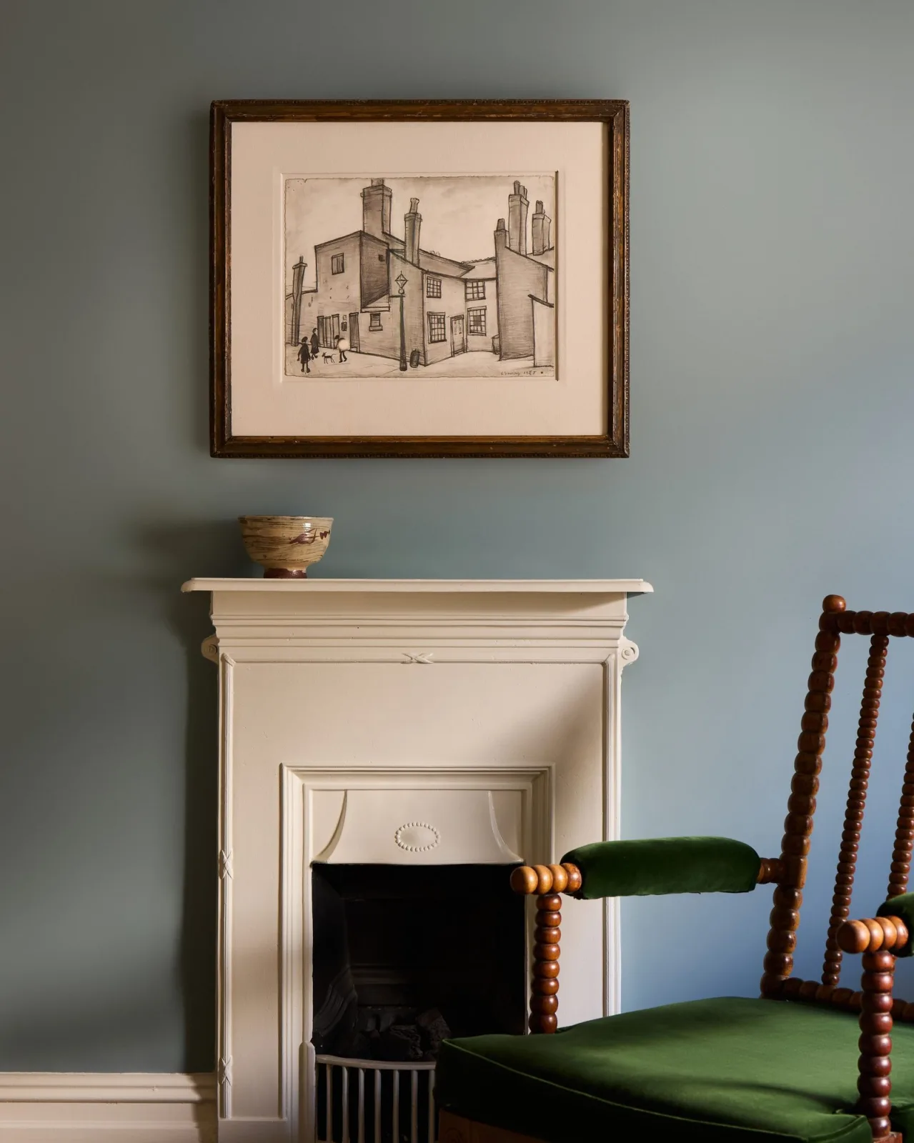 Framed pencil drawing of a row of terraced houses with tall chimneys and a few small figures and a dog in the street, hanging on a blue-grey wall above a cream fireplace with a green velvet armchair beside it. On view at Jenna Burlingham Gallery.