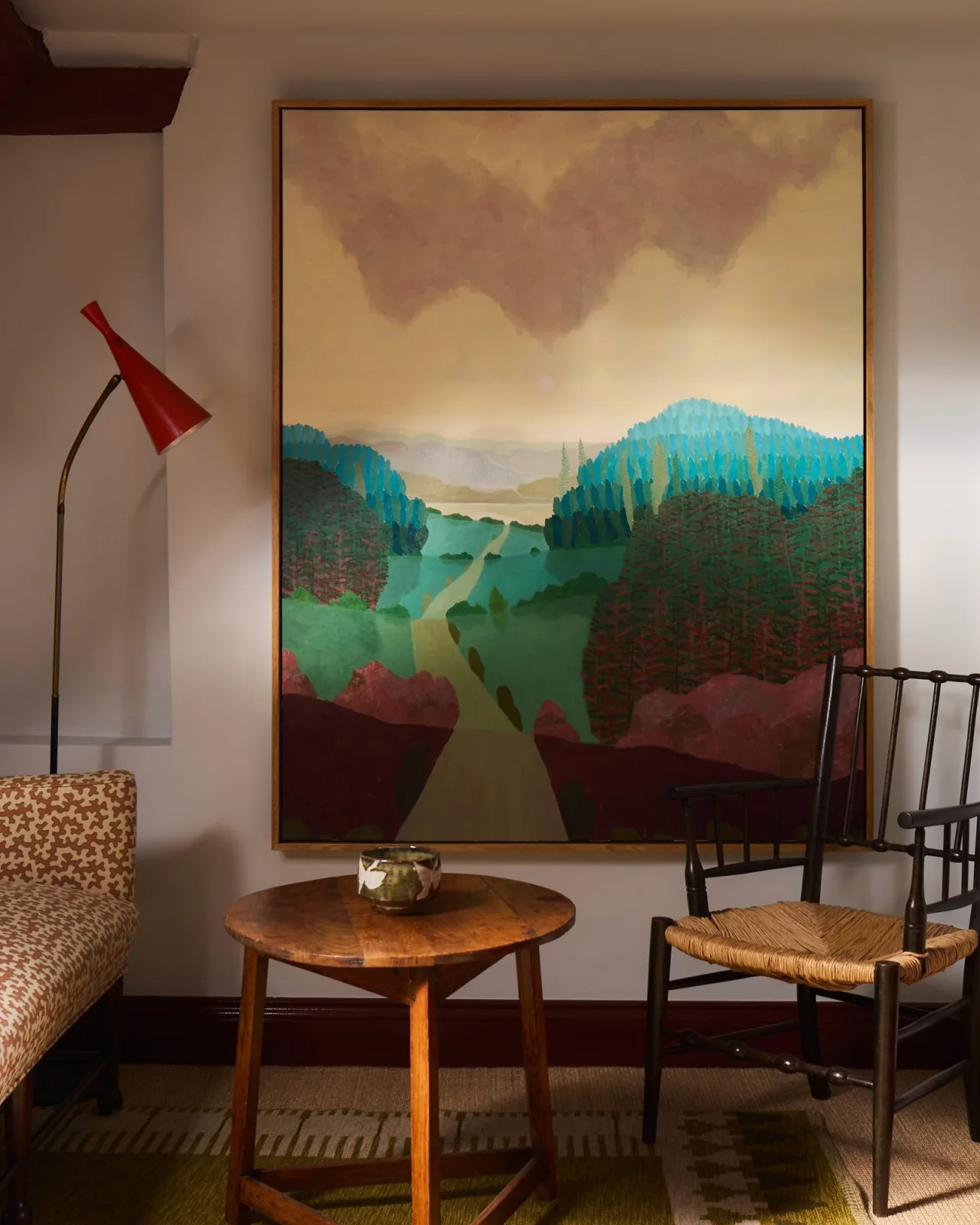 Interior view of a sitting room featuring a large painting by Richard Ballinger titled “From the Red to the Blue” (2026), depicting a stylized landscape with a winding path leading through forested hills in teal, green, and deep red tones beneath a hazy, mauve-clouded sky. The large wood-framed painting hangs above a round wooden side table holding a ceramic bowl, flanked by a woven-seat chair and a patterned sofa, with a red cone-shaped floor lamp to the left. On view at Jenna Burlingham Gallery in the UK.