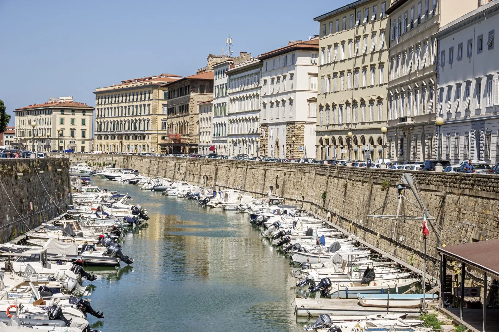 a canal with various sail boats anchored below a row of houses with small windows on a bright sunny day